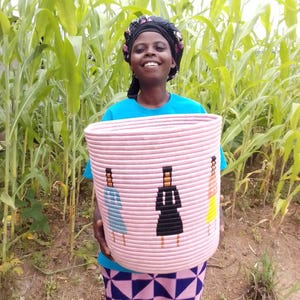 May include: A pink woven basket with three stylized figures in blue, black, and yellow. The basket is cylindrical and appears to be handmade. The basket is held by a person wearing a blue shirt and a patterned skirt.
