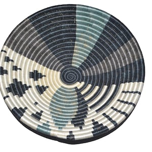 May include: A circular woven basket with a spiral pattern. The basket has alternating sections of white, black, and teal. The design incorporates geometric shapes and concentric circles, creating a visually appealing pattern.