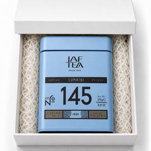 Op de afbeelding: Een lichtblauw rechthoekig theeblik met het JAF TEA logo, de tekst Single Lumbini Estate, Lot No 145 en een nettogewicht van 175g. Het blik wordt gepresenteerd in een witte doos met wit beschermend opvulmateriaal van versnipperd papier.
