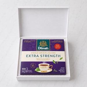 Puede incluir: Una caja blanca abierta que contiene un paquete cuadrado de té negro Dilmah Extra Strength Single Origin Ceylon. El envase morado y crema muestra un gráfico de una taza de té, el texto 100 bolsitas de té y un peso neto de 240g.