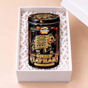 Puede incluir: Una lata de té cilíndrica negra con una ilustración de elefante dorada y colorida, el texto Battler, Big Leaf OP, Pure Ceylon Tea, Black Elephant y texto cirílico. La lata está dentro de una caja blanca con relleno de papel blanco.