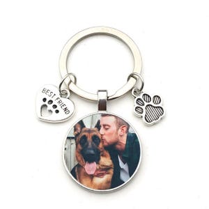 May include: A silver keyring with a round pendant showing a photo of a person kissing a German Shepherd dog. The keyring includes a heart charm with the text "BEST FRIEND" and a paw print charm.