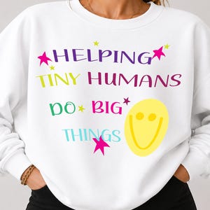 Può includere: Una felpa bianca a girocollo con il testo multicolore HELPING TINY HUMANS DO BIG THINGS. Il design include una grande faccina sorridente gialla e diverse piccole stelle rosa e gialle sparse attorno alla scritta sulla parte anteriore.