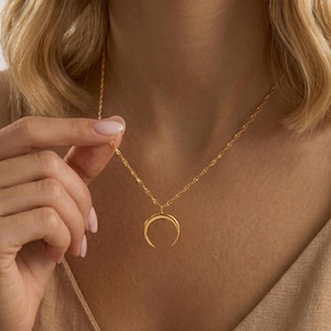 May include: A gold-toned necklace featuring a delicate twisted chain and a smooth, polished crescent moon pendant. The pendant hangs centrally from the chain, which rests against the collarbone. The metal has a bright, reflective finish.