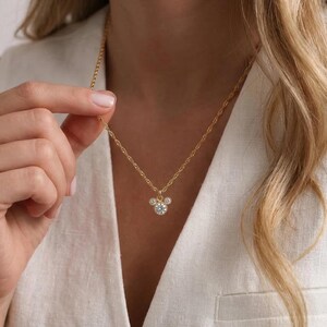 Peut inclure: Un collier doré avec un pendentif en forme de tête de souris. Le pendentif est orné de pierres claires et scintillantes. Le collier est tenu par une personne portant un blazer blanc.