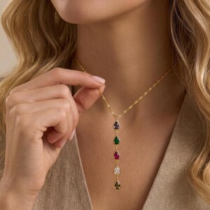 May include: A gold-colored necklace with a delicate chain and a vertical arrangement of five teardrop-shaped pendants. Each pendant features a different colored gemstone: purple, green, red, white, and olive green. The necklace is being held up by a person.
