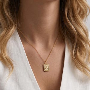 May include: Gold-colored necklace with a rectangular pendant featuring the letter "A". The pendant has a textured border and is suspended from a delicate chain. The necklace is worn against a white top.