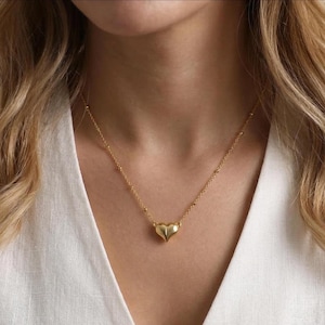 Könnte beinhalten: Eine zarte goldene Halskette mit einem herzförmigen Anhänger. Die Kette hat eine feine Gliederkette mit kleinen, gleichmäßig verteilten Perlen. Der Herzanhänger ist eine solide, dreidimensionale Form mit glatter, polierter Oberfläche. Die Halskette wird über einem weißen Oberteil getragen.