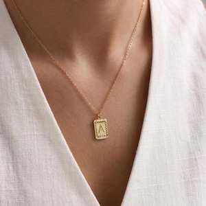 Può includere: Collana color oro con un ciondolo rettangolare con la lettera "A". Il ciondolo ha un bordo strutturato e pende da una delicata catena. La collana è indossata su un top bianco.