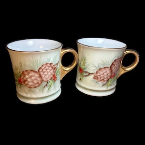 May include: Two porcelain mugs featuring a painted design of brown pinecones, green pine needles, and small red berries. Each mug has a white interior, a cream-colored exterior, and a metallic gold handle with a matching gold rim.