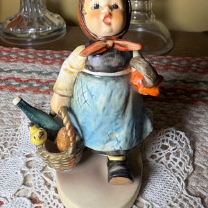 May include: A ceramic figurine of a person wearing a headscarf, a dark bodice, and a blue apron. The figure carries a woven basket containing a green bottle, a yellow bird, and a brown loaf, while holding a small orange object in the other hand.