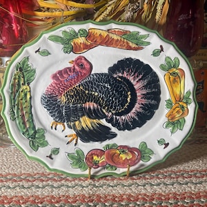 Puede incluir: Una bandeja de servicio ovalada de cerámica con borde festoneado verde, que presenta un pavo pintado a mano en el centro con plumaje negro, amarillo y rojo. El borde está decorado con ilustraciones de zanahorias, pepinos, pimientos amarillos y tomates rojos.