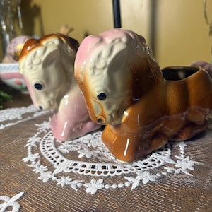 Könnte beinhalten: Zwei Keramikpferdefiguren. Eine ist rosa und cremefarben, die andere braun und cremefarben. Beide haben schwarze Augen und stehen auf einer weißen Spitzendecke. Die Pferdefiguren sind wahrscheinlich dekorative Vintage- oder Antiquitäten.