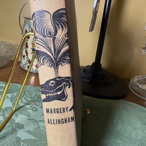 Puede incluir: Un libro antiguo de tapa dura titulado "Black Plumes" de Margery Allingham. El libro tiene una cubierta de color beige con elementos decorativos y texto en negro. La columna vertebral presenta una ilustración estilizada y el logotipo "Sun Dial Mysteries".