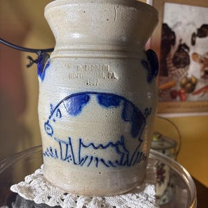 May include: A vintage stoneware crock with a blue pig design. The off-white crock has two small blue handles and text that reads "D. Fiedrich, Northhampton, PA. 1957". The crock is sitting on a glass plate with a white lace doily.