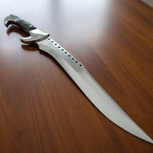 May include: A silver and black dagger with a curved blade and a decorative handle. The blade has a series of small holes along the spine. The dagger is lying on a wooden surface.