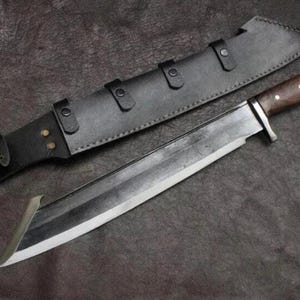 May include: A large fixed-blade knife with a dark, polished steel blade featuring a distinct angled tip. The handle is crafted from dark brown wood with three silver rivets. It is accompanied by a black leather sheath with metal rivets.