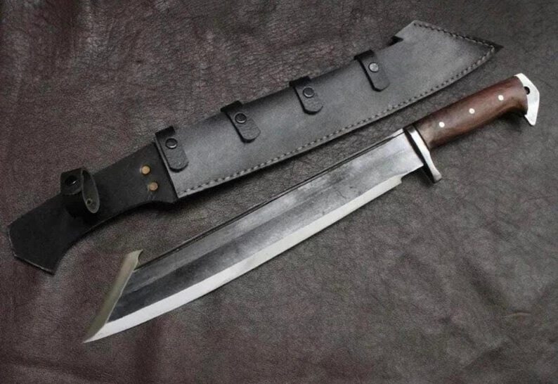 May include: A large fixed-blade knife with a dark, polished steel blade featuring a distinct angled tip. The handle is crafted from dark brown wood with three silver rivets. It is accompanied by a black leather sheath with metal rivets.