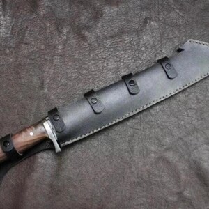 May include: A fixed-blade knife with a polished wooden handle and a silver-toned bolster, secured within a black leather sheath. The sheath features visible stitching along the edges and three black leather straps with metal snaps for closure.