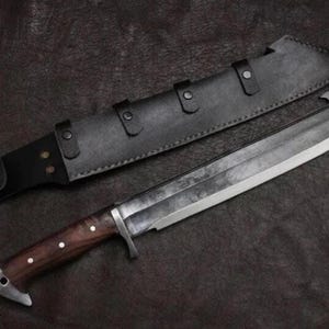 May include: A large fixed-blade knife with a dark, textured steel blade and a polished wooden handle secured with three metal rivets. The knife features a metal crossguard and a pommel, accompanied by a black leather sheath with metal snap closures.