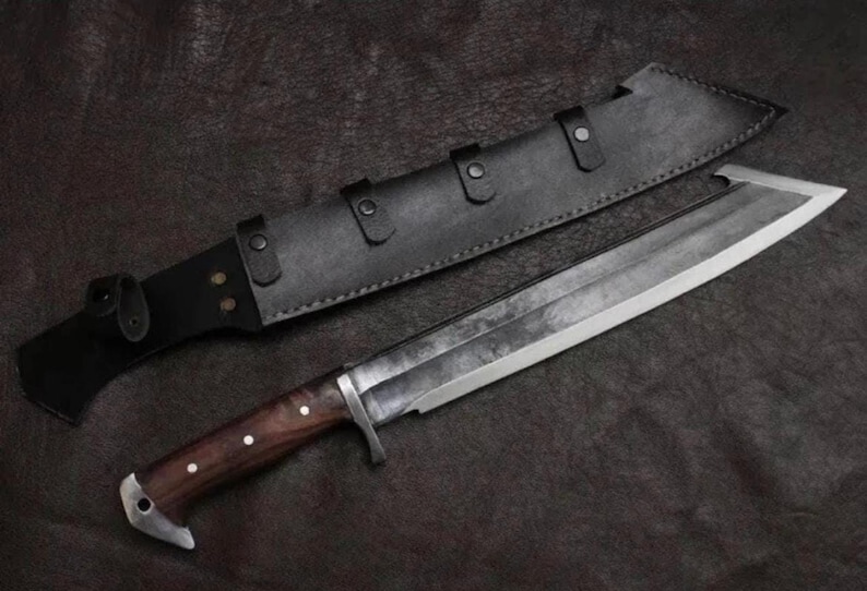 May include: A large fixed-blade knife with a dark, textured steel blade and a polished wooden handle secured with three metal rivets. The knife features a metal crossguard and a pommel, accompanied by a black leather sheath with metal snap closures.