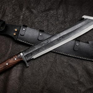 May include: A fixed-blade knife with a dark, textured steel blade and a polished wooden handle featuring three silver pins. The knife includes a black leather sheath with metal rivets and a strap closure, designed for secure storage and transport.