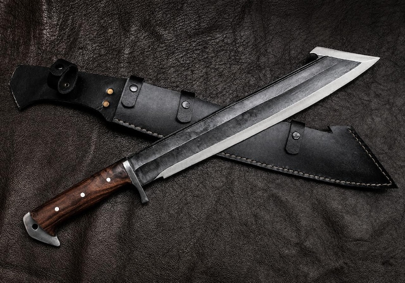 May include: A fixed-blade knife with a dark, textured steel blade and a polished wooden handle featuring three silver pins. The knife includes a black leather sheath with metal rivets and a strap closure, designed for secure storage and transport.
