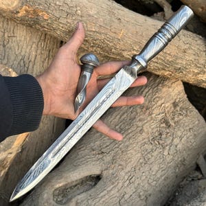 Può includere: Un pugnale artigianale in acciaio di Damasco con un tappo abbinato. Il pugnale presenta una lama lunga e appuntita e un'impugnatura strutturata. Il tappo della bottiglia ha un design a spirale. Entrambi gli articoli hanno un aspetto unico e modellato.