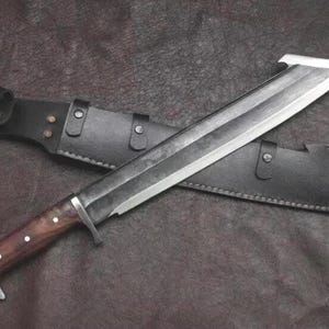 May include: A large fixed-blade knife with a dark, weathered steel blade featuring a hooked tip, a polished metal bolster, and a smooth brown wooden handle with three silver rivets. The knife is accompanied by a black leather sheath with metal studs.