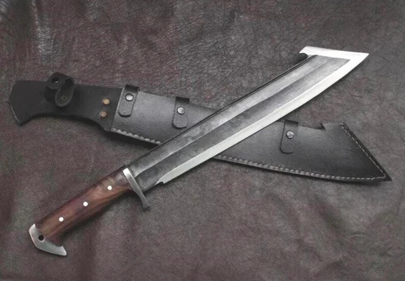 May include: A large fixed-blade knife with a dark, weathered steel blade featuring a hooked tip, a polished metal bolster, and a smooth brown wooden handle with three silver rivets. The knife is accompanied by a black leather sheath with metal studs.
