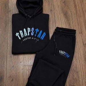 May include: A black hooded sweatshirt and matching sweatpants set. The hoodie features the text TRAPSTAR in a blue gradient font with the text IT'S A SECRET underneath. The sweatpants feature the same TRAPSTAR logo in blue on the upper left leg.