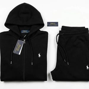 Puede incluir: Conjunto de chándal negro compuesto por una sudadera con capucha y cremallera y un pantalón de chándal a juego. La sudadera tiene una capucha con cordón y un pequeño logotipo blanco. El pantalón de chándal también tiene un logotipo blanco y cintura con cordón.