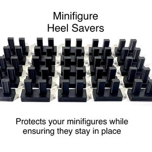 May include: Black plastic Minifigure Heel Savers. The image shows several sets of black plastic stands designed to protect and secure miniature figures. The text reads "Protects your minifigures while ensuring they stay in place."