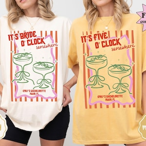 May include: Ivory and mustard-colored t-shirts with a graphic design. The design features cocktail glasses, the text "IT'S BRIDE O'CLOCK somewhere" and "IT'S FIVE O'CLOCK somewhere". The text "EMILY'S BACHELORETTE MIAMI FL" is also included.