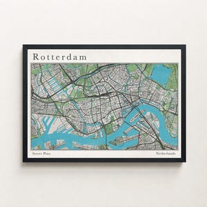 Può includere: Una mappa stradale incorniciata di Rotterdam, Paesi Bassi. La mappa presenta un'illustrazione dettagliata della disposizione della città, con corsi d'acqua blu, parchi verdi e strade grigie. La parola "Rotterdam" è stampata in alto e "Street Plan" e "Netherlands" in basso.