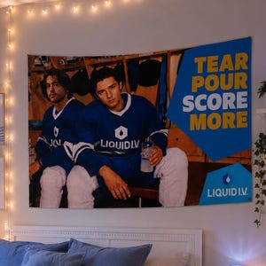 May include: A large rectangular wall tapestry featuring two individuals in blue hockey jerseys with the text "TEAR POUR SCORE MORE" and "LIQUID I.V." displayed prominently. The tapestry is hung above a bed with blue bedding and a white knit throw blanket.