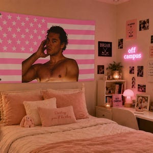 May include: A pink and white American flag tapestry with a shirtless man's image hangs above a bed with pink bedding. The room is decorated with photos, a neon sign that says "off campus", and a small desk.