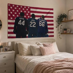 May include: A rectangular wall tapestry featuring a red, white, and blue American flag background with three hockey players in dark jerseys standing with their backs to the viewer, displaying the names Graham 44, Logan 22, and Di Laurentis 66.