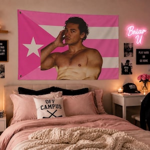 May include: A rectangular wall tapestry featuring a pink and white horizontal striped design with a white star on a pink triangle on the left side. The center of the tapestry displays a photographic print of a shirtless person with dark hair.