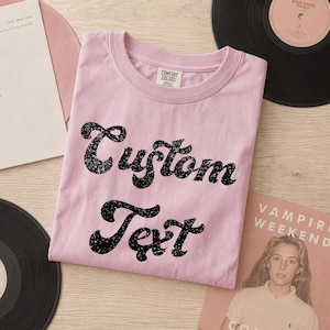 Puede incluir: Una camiseta de manga corta de color rosa claro con cuello redondo y una etiqueta de Comfort Colors en el interior del cuello. El frente presenta el texto negro de estilo desgastado Custom Text en una fuente cursiva negrita.