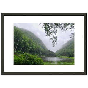 May include: A framed landscape photograph featuring a serene lake nestled between lush, green mountains. The image is framed in a dark gray border, with a white matting. The sky is overcast, adding a soft, diffused light to the scene.