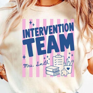 May include: An ivory t-shirt featuring a graphic with vertical pink stripes and the blue text "INTERVENTION TEAM" above "Mrs. Smith". The design includes blue illustrations of a clipboard, two stacked books, a pencil holder, and a puzzle piece.