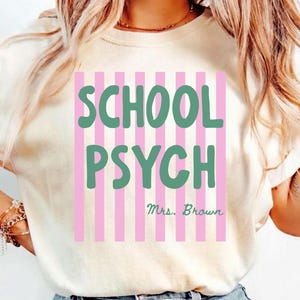 May include: An ivory short-sleeved t-shirt featuring a graphic design with vertical pink stripes. The text SCHOOL PSYCH is printed in large green letters, with the name Mrs. Brown written in smaller green cursive script underneath the text.