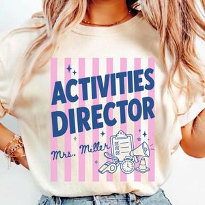 May include: An ivory t-shirt featuring a graphic with vertical pink stripes and the dark blue text "ACTIVITIES DIRECTOR" above "Mrs. Miller". The design includes a clipboard, a whistle, a megaphone, a stopwatch, and a traffic cone icon.