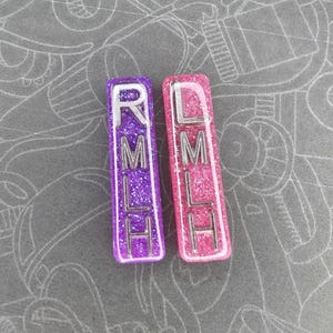 May include: Two rectangular resin charms, one purple and one pink, with silver lettering spelling out "RMLH" and "LMLH". The charms have glitter embedded in the resin. The background is a gray surface with a white pattern.