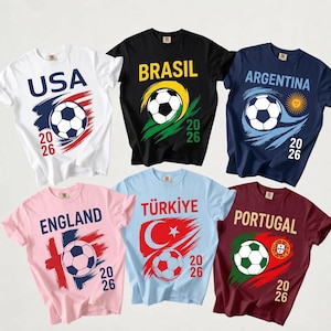 Può includere: Sei magliette a maniche corte in bianco, nero, blu navy, rosa chiaro, azzurro e bordeaux, ognuna con una grafica di un pallone da calcio, l'anno 2026 e i nomi USA, BRASIL, ARGENTINA, ENGLAND, TÜRKIYE e PORTUGAL con motivi nazionali.