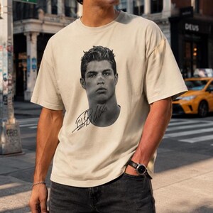 May include: A beige short-sleeved t-shirt featuring a black and white portrait of a man with short, styled hair. Below the portrait is a black handwritten-style signature. The shirt has a crew neckline and a relaxed, loose-fitting silhouette.
