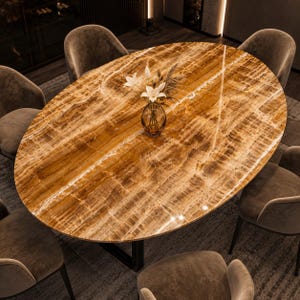 Puede incluir: Una mesa de comedor ovalada con una superficie de piedra pulida color ámbar que presenta vetas blancas naturales. La mesa está rodeada por seis sillas tapizadas en gris y sostiene un jarrón de vidrio transparente con flores blancas y hierbas.
