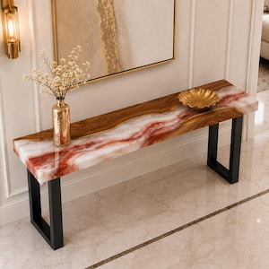 Puede incluir: Una mesa consola rectangular que presenta un tablero de madera de borde natural combinado con una incrustación de resina pulida con efecto mármol rojo y blanco. La mesa se apoya en dos patas de metal negro en forma de U y tiene un jarrón dorado.