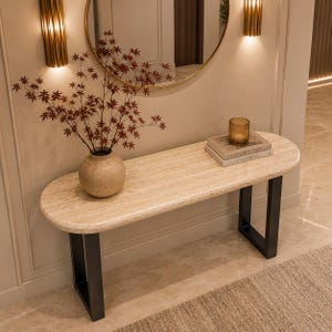 Puede incluir: Una mesa consola con una superficie ovalada de travertino beige liso, sostenida por dos patas de metal negro en forma de U. Un jarrón de cerámica redondo con ramas de arce rojo se sitúa sobre la mesa junto a dos libros y un portavelas de cristal.